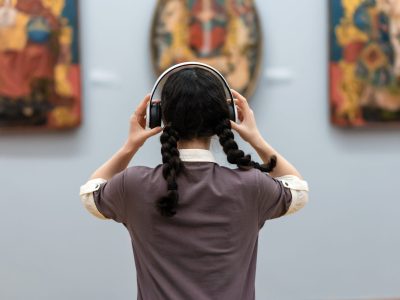 Back view of young woman wearing headphones and contemplates ancient arts. Student visiting gallery or museum. Concept of modern education and culture.